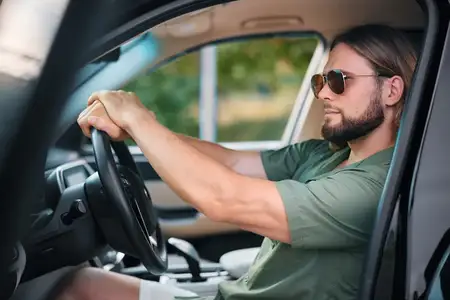 Стильный молодой человек в солнцезащитных очках сидит в автомобиле, уверенно держась за руль