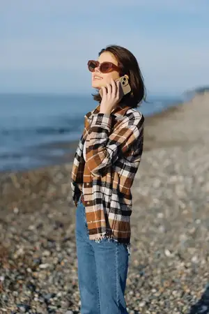 Непринужденная прибрежная сцена: женщина в клетчатой рубашке и джинсах разговаривает по мобильному телефону у воды
