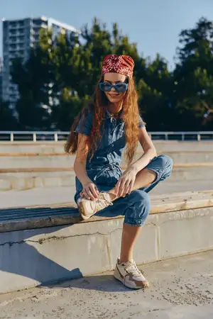 Женщина в повседневном образе, одетая в современный бохо-вестерн деним в стиле стрит-стайл, сидит на бетонных ступеньках в солнцезащитных очках и бандане, наслаждаясь солнечной городской обстановкой…