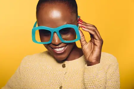 Счастливая темнокожая африканка поправляет большие синие солнцезащитные очки, одетая в уютный жёлтый свитер