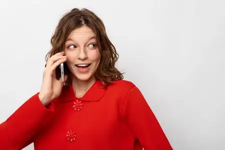 Улыбающаяся женщина в красном трикотаже прижимает телефон к уху и смотрит в сторону с удивленным лицом
