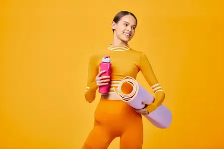 Спортивная девушка в яркой одежде с ковриком для йоги и бутылкой демонстрирует энергичную фитнес-программу, атлетические движения и здоровый образ жизни в смелой студийной обстановке с яркими цветами