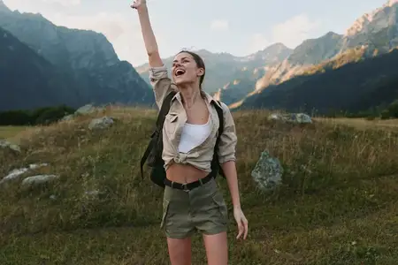 Улыбающаяся молодая женщина в белом топе, хаки-шортах и лёгкой куртке стоит в открытой долине на фоне возвышающихся вершин, излучая уверенность и дух приключений в естественной обстановке