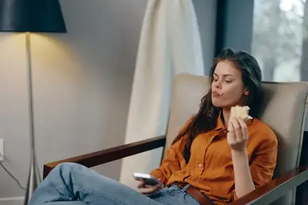 Молодая женщина удобно сидит в кресле, ест полезную закуску и смотрит телевизор