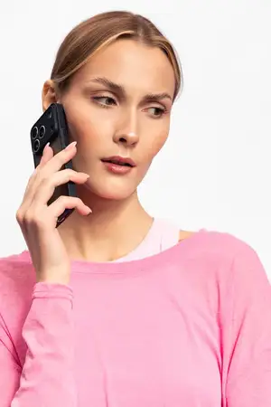 Серьезная молодая женщина в розовом свитере держит смартфон у уха, глядя в сторону на белом фоне