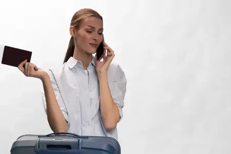 Женщина с паспортом и смартфоном стоит рядом с чемоданом, разговаривая по телефону
