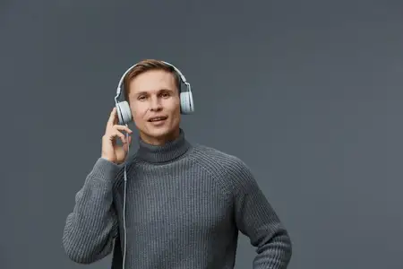 Молодой мужчина европеоидной расы в сером вязаном свитере и белых наушниках смотрит в камеру и взаимодействует с голосовым помощником для управления и автоматизации умного дома на темно-сером фоне…