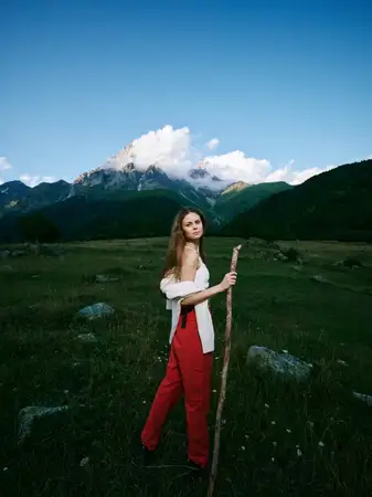 Уличная сцена: женщина с посохом на лугу в долине, вдали горы и ясное небо