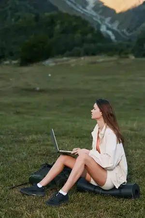 Спокойная женщина-пользователь работает с ноутбуком, сидя на траве, на фоне холмов и голубого неба, что передает продуктивность и концентрацию на открытом воздухе