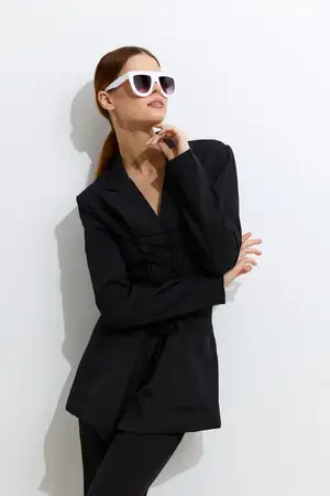 Красивая женщина в черном tailored блейзере и белых солнцезащитных очках позирует с рукой у лица, излучая модное очарование и уверенность в яркой минималистичной студии