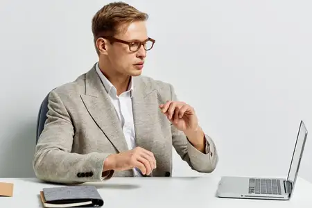 Уверенный профессиональный мужчина в стильном сером пиджаке и белой рубашке работает за ноутбуком за столом в чистом светлом офисном помещении, анализируя данные или готовя презентацию