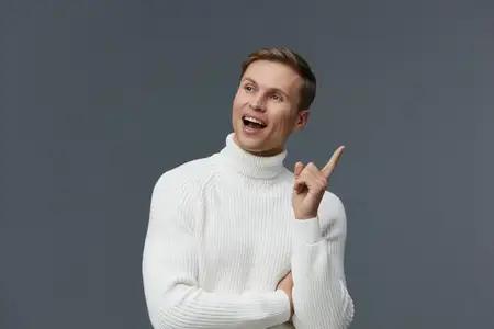 Улыбающийся мужчина европеоидной расы в позе мыслителя с поднятым пальцем, одетый в белую вязаную водолазку, изолированный на сером фоне, демонстрирующий позитивные эмоции и взаимодействие