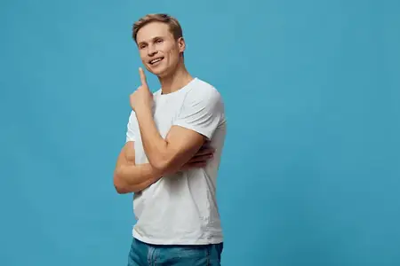 Счастливый молодой человек со светлыми волосами в белой футболке свободного кроя стоит на синем фоне