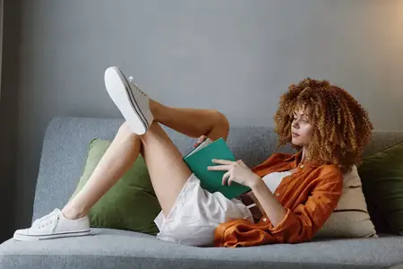 Модная молодая женщина с вьющимися волосами с удовольствием читает книгу, развалившись на мягком диване