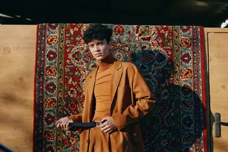 Модный молодой человек уверенно позирует с пистолетом на фоне яркого, замысловатого гобелена