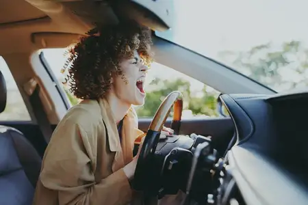В солнечном салоне автомобиля она сжимает рулевое колесо и наклоняется вперед, одетая в бежевый кардиган, который сочетается с теплой ретро-цветовой палитрой, создавая яркое, энергичное настроение