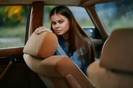 Безмятежная молодая женщина с длинными каштановыми волосами в джинсовой куртке позирует в салоне автомобиля на фоне природы