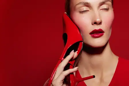 Портрет женщины с закрытыми глазами и глянцевыми красными губами, держащей красную туфлю-шпильку возле щеки