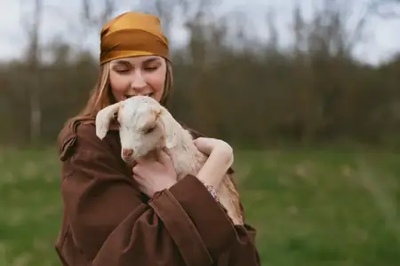 Молодая женщина в коричневом пальто и оранжевом платке крепко держит на руках белого козлёнка, стоя на зелёном поле с размытыми деревьями на заднем плане в пасмурный день