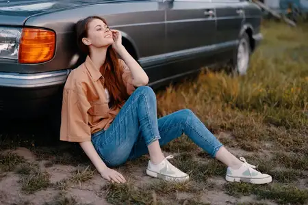 Молодая модель с длинными волосами сидит рядом с винтажным автомобилем в солнечный день, одетая в повседневную одежду, чувствуя себя расслабленно в естественной обстановке, что передает беззаботный…