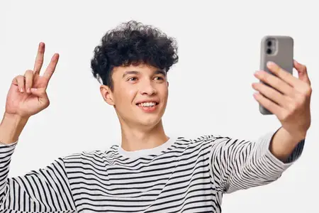 Улыбающийся мужчина делает селфи на смартфон в повседневной полосатой рубашке, показывая жест «мир» на фоне чистого белого фона, выражая радость и уверенность