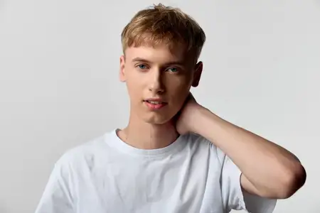 Повседневная мужская модель в белой футболке с рукой на шее на однотонном фоне, демонстрирующая спокойное выражение лица и современный стиль для рекламы, редакционных материалов или модельных…