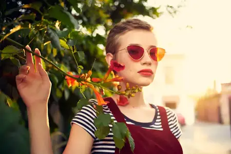 Женщина в солнцезащитных очках стоит рядом с кустом цветов на улице, ведёт активный образ жизни