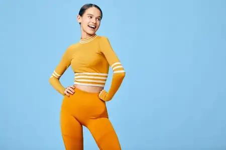 Спортивная женщина-модель в ярком оранжевом спортивном костюме с укороченным топом и штанами с высокой талией застыла в динамичной позе, подчеркивающей энергию фитнеса, баланс и решимость в чистом…