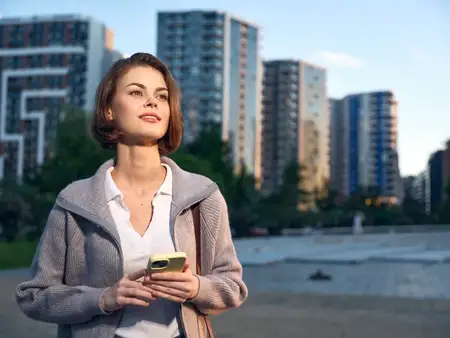 Городская профессиональная женщина смотрит вдаль, держа телефон, излучая спокойную уравновешенность и амбициозность на фоне современного города