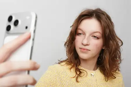 Женщина с вьющимися волосами в желтом свитере держит смартфон для селфи на однотонном фоне