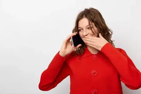 Улыбающаяся женщина в красном джемпере держит телефон у уха и прикрывает рот рукой, выражая удивление