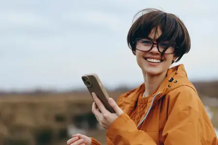 Портрет улыбающейся женщины на открытом воздухе в оранжевой куртке, использующей смартфон, расслабленная поза, открытое небо на заднем плане, путешествие, общение и цифровая жизнь, свежий воздух…