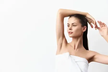 Выразительное изображение модели с длинными волосами и гладкой кожей, которая поднимает руки, будучи завёрнутой в белое полотенце, подчёркивая уверенность и грацию