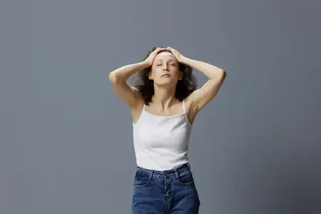 Уставшая, расслабляющаяся, спокойная, кудрявая, красивая женщина в простой белой футболке наслаждается массажем головы, закрыв глаза, позирует изолированно на серо-голубом фоне