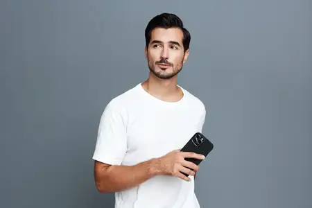 Задумчивый молодой человек в повседневной белой футболке держит смартфон и задумчиво смотрит в сторону на фоне минималистичного серого фона