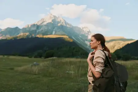 Женщина-туристка с рюкзаком наслаждается горным видом в солнечную погоду, стоя на травянистом поле, мирная природная сцена, активный образ жизни, концепция путешествия