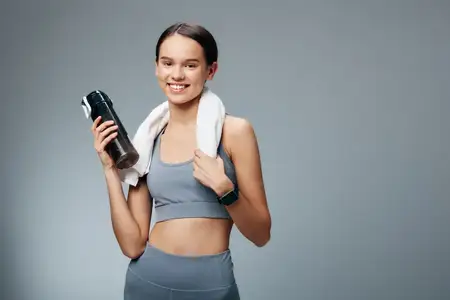 Спортивная женщина-атлет уверенно позирует в спортивной одежде с умными часами и видимым подтянутым прессом на нейтральном сером фоне, подчёркивая режим упражнений, восстановление после тренировки и…