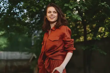Молодая женщина стоит в лиственной обстановке на открытом воздухе в ржавом комбинезоне, тепло улыбается, а мягкий солнечный свет подчеркивает ее расслабленное и уверенное настроение