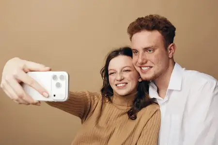 Счастливая молодая пара улыбается, делая селфи, женщина в коричневом свитере и мужчина в белой рубашке близко друг к другу на бежевом фоне