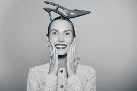 Счастливая светлокожая женщина-модель европейской внешности балансирует туфлей на высоком каблуке на голове, держа руки у лица, одетая в трикотаж