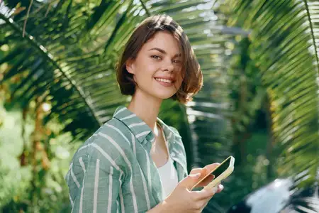 Расслабленная уличная сцена, запечатлевшая повседневную моду, естественный свет и повседневные технологии в пышной зеленой обстановке