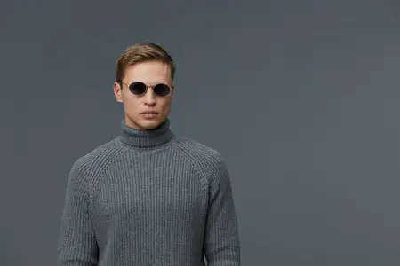 Утончённый мужчина с короткими волосами и солнцезащитными очками, одетый в серую водолазку, позирует на простом тёмном фоне, излучая уверенность и стиль