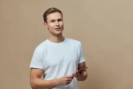 Улыбающийся молодой мужчина европеоидной внешности держит смартфон, пользуется мобильным телефоном в повседневной белой футболке на бежевом фоне