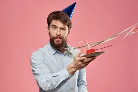 Бородатый мужчина с тортом и высунутым языком на розовом фоне, обрезной вид, на голове синий колпак