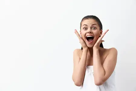 Радостная молодая женщина с длинными тёмными волосами, одетая в полотенце, выражает восторг и удивление: широко открытые глаза и улыбка с приоткрытым ртом