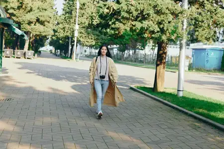 Повседневный шик в движении: женщина в длинном пальто и джинсовой одежде прогуливается по городскому тротуару, атмосфера городского парка с солнечным светом и деревьями, уверенный момент уличной моды