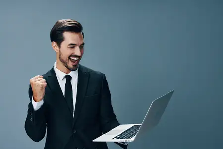 Мужчина в деловом костюме смотрит в ноутбук и работает онлайн, улыбаясь и поднимая сжатый кулак вверх, что символизирует победу