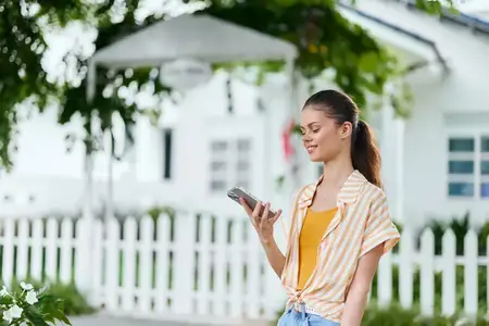 Веселая молодая женщина стоит в ярком саду, увлеченно глядя в смартфон