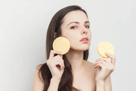Брюнетка с обнаженными плечами вытирает лицо губкой, чистая кожа