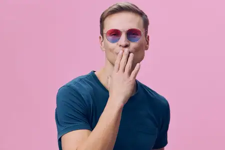 Молодой человек посылает воздушный поцелуй, улыбается, одет в синюю футболку, розовые и синие солнцезащитные очки, изолирован на розовом фоне